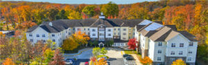 Aerial Exterior Victoria Park at Edgewater, Residential building with parking lot in the front, meticulous landscaping, lush fall foliage, sprawling hills in the background, photo taken on a sunny day with clouds.