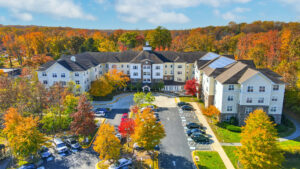 Aerial Exterior Victoria Park at Edgewater, Residential building with parking lot in the front, meticulous landscaping, lush fall foliage, sprawling hills in the background, photo taken on a sunny day with clouds.