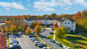 Aerial Exterior Victoria Park at Edgewater, Residential building with parking lot in the front, meticulous landscaping, lush fall foliage, sprawling hills in the background, photo taken on a sunny day with clouds.