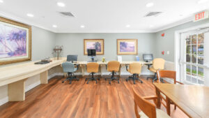 Interior Victoria Park at Edgewater Computer Room, Wood floors, 5 computer stations with 3 desktops, olive walls, landscape art on the walls, emergency exit in room.
